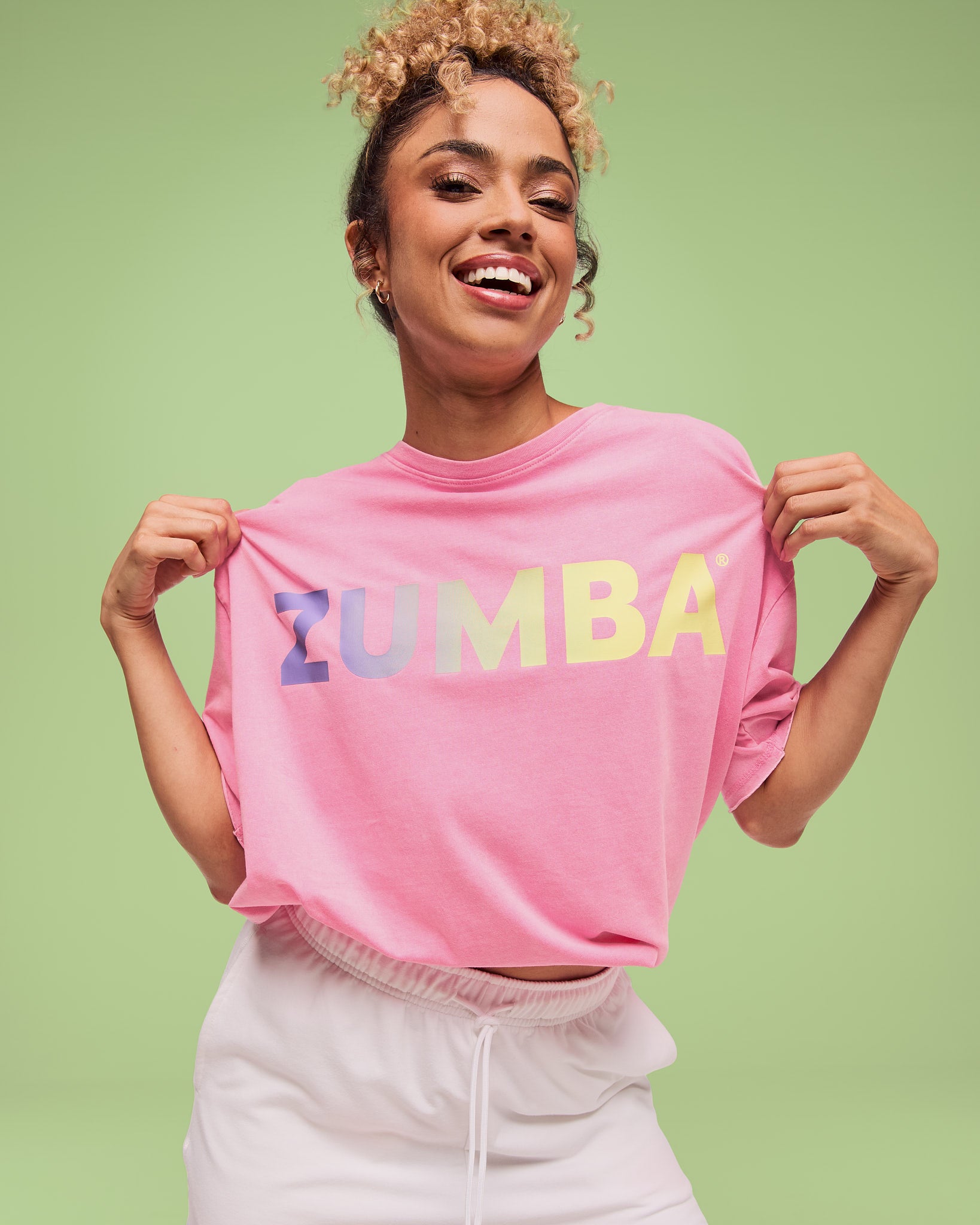 Female model wearing a pink short-sleeve T-shirt with a pastel gradient “ZUMBA” logo across the chest, paired with white drawstring shorts.