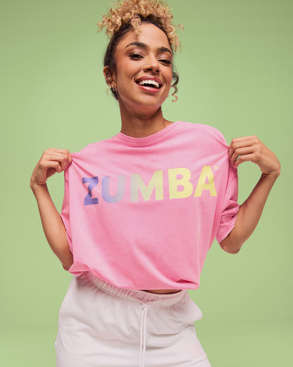 Female model wearing a pink short-sleeve T-shirt with a pastel gradient “ZUMBA” logo across the chest, paired with white drawstring shorts.