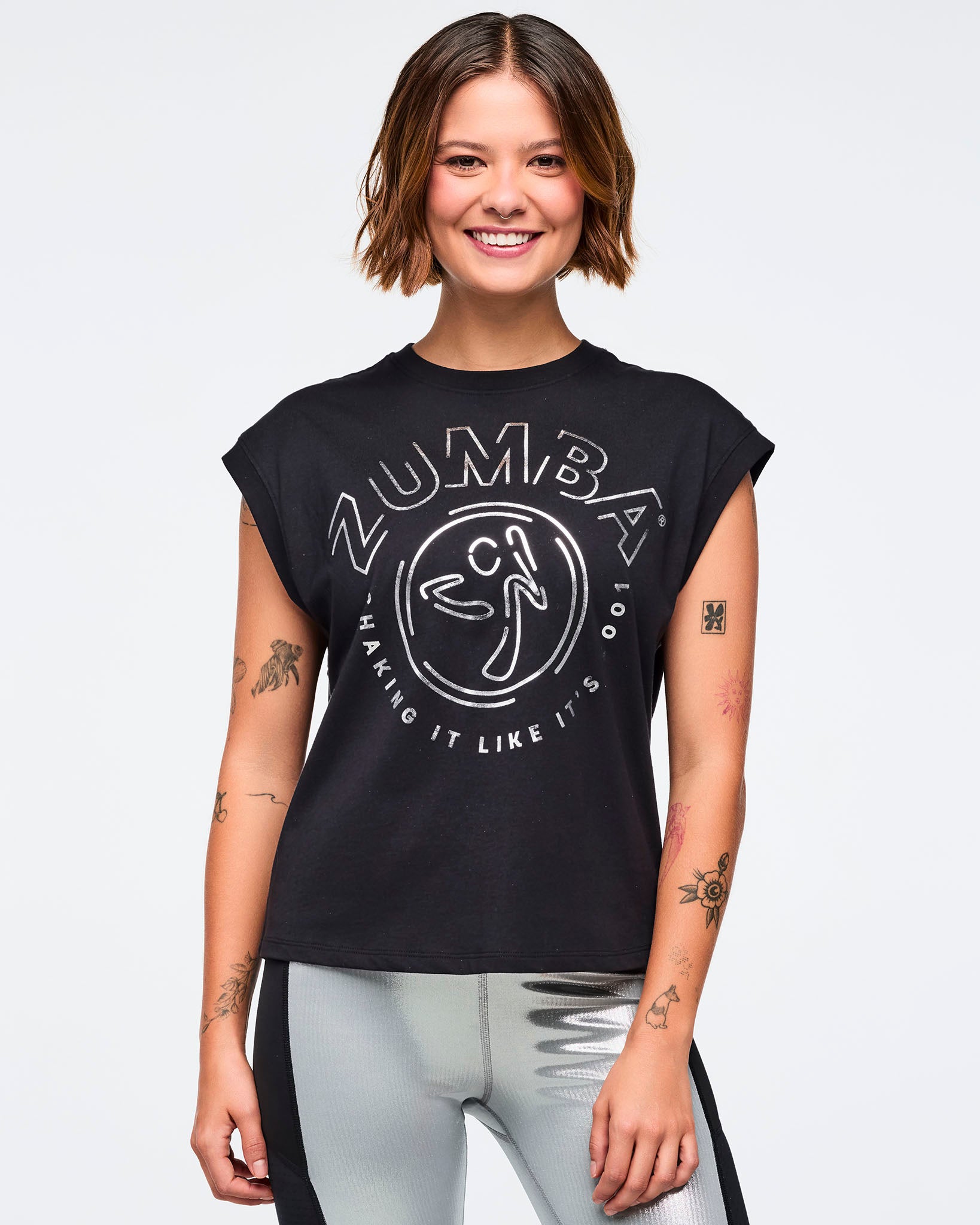 Black cap-sleeve Zumba graphic top with a large silver logo print on the front. Female model wears the relaxed-fit top with metallic silver leggings featuring black side panels, smiling in a front view against a light gray background.
