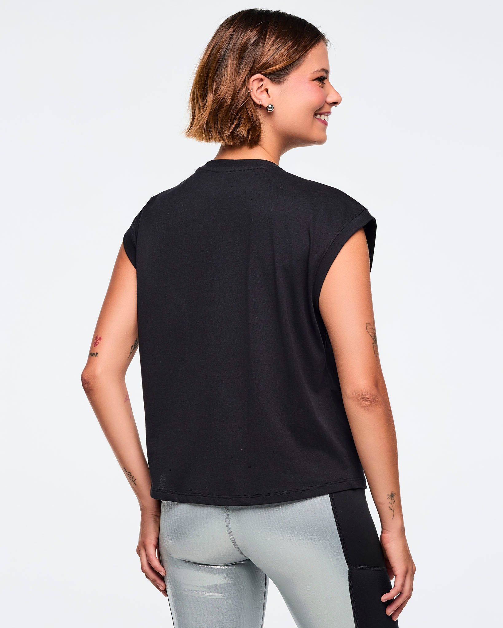 Black cap-sleeve Zumba graphic top with a plain solid back. Female model wears the relaxed-fit top with metallic silver leggings featuring black side panels, shown from the back against a light gray background.