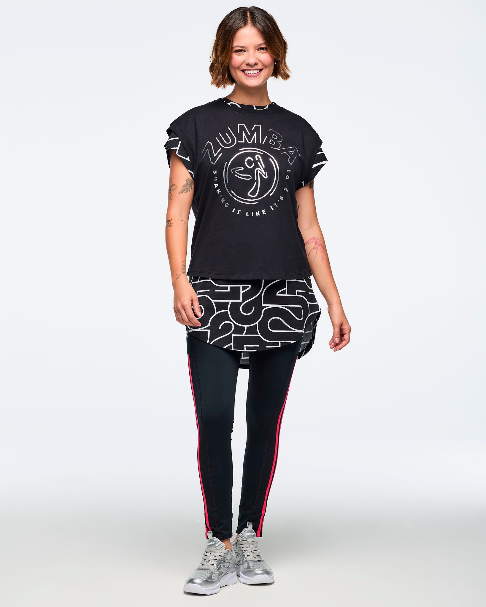 Black cap-sleeve Zumba graphic top with a large silver front logo. Female model wears the top layered over a long black-and-silver patterned shirt, black leggings with bright red side stripes, and silver athletic sneakers, standing in a full-body front view against a light gray background.