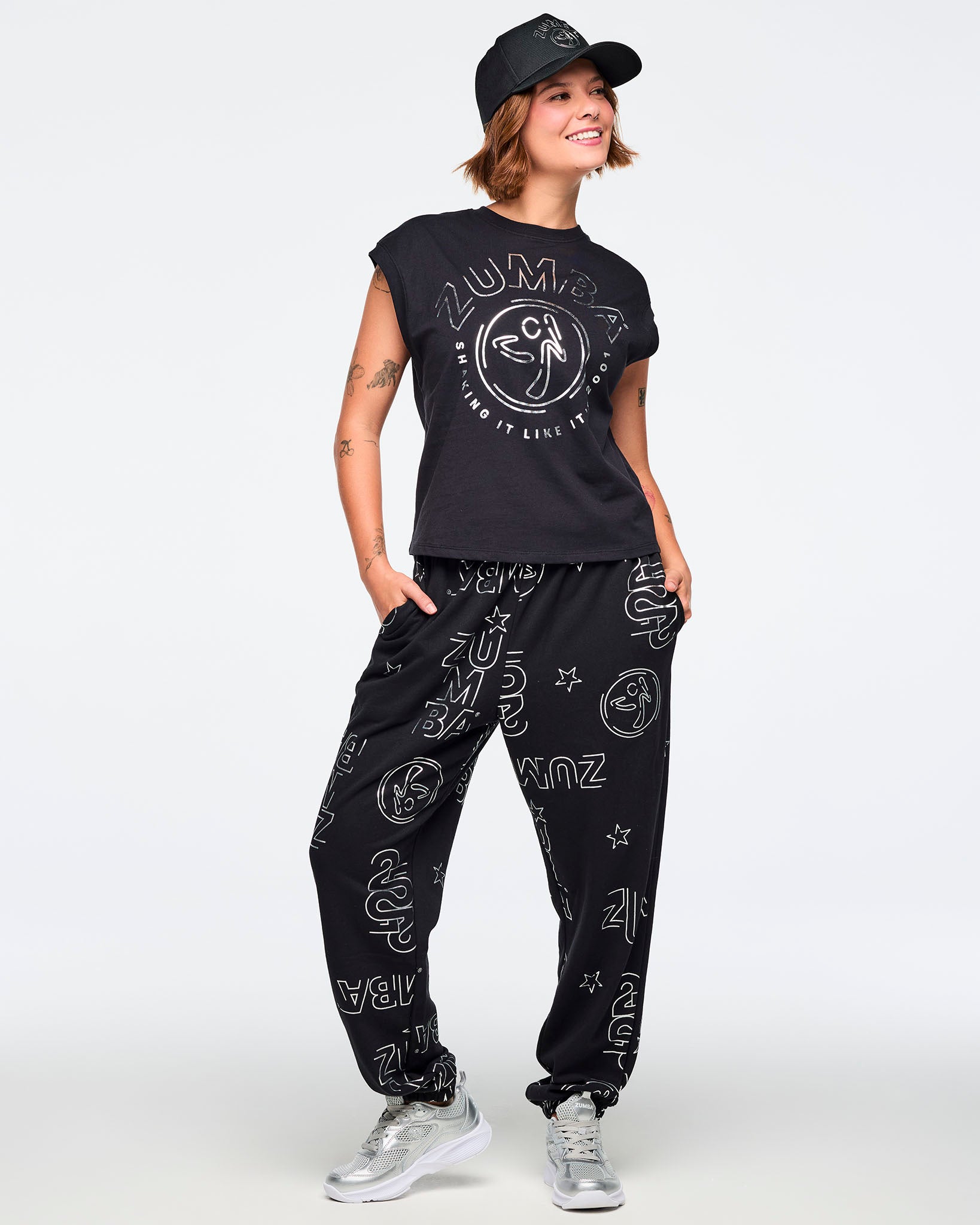Black cap-sleeve Zumba graphic top with a large silver front logo. Female model wears the relaxed-fit top with black jogger pants covered in silver Zumba prints, a black baseball cap, and silver athletic sneakers, posing against a light gray background.