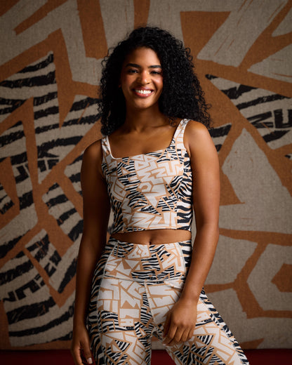 Female model wearing a fitted cropped tank in a beige, cream, and black abstract zebra-print pattern with a square neckline and wide shoulder straps, paired with matching high-waisted leggings against a colorful background.