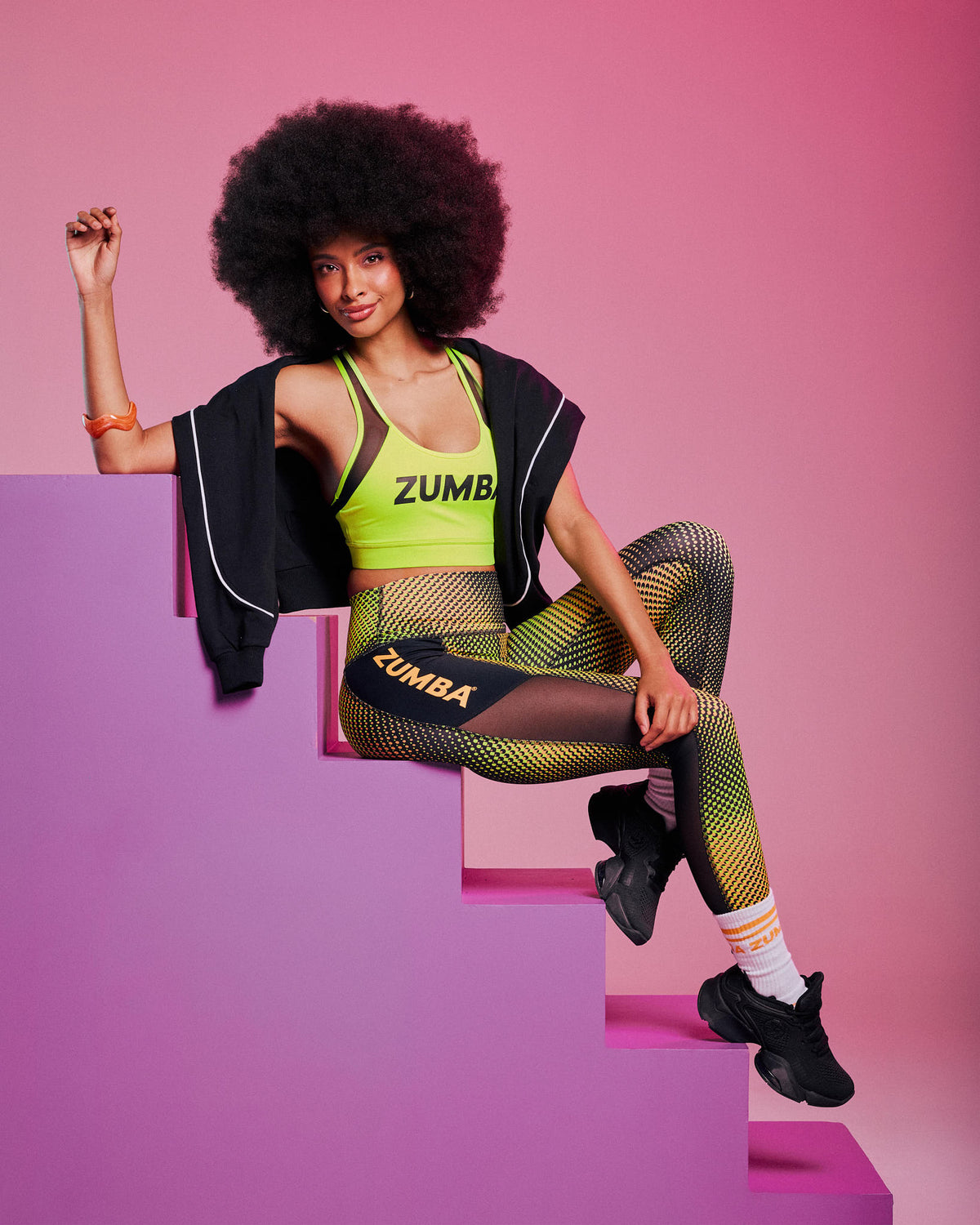 Female model with an afro seated on pink steps, wearing a yellow-green "ZUMBA" sports bra, matching gradient leggings with "ZUMBA" text, black sneakers, white socks, and a black jacket draped off her shoulders, against a pink background.