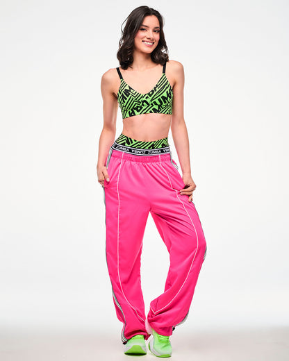 Full view of a female model wearing a neon green and black geometric striped print bra, paired with pink track pants and green shoes.
