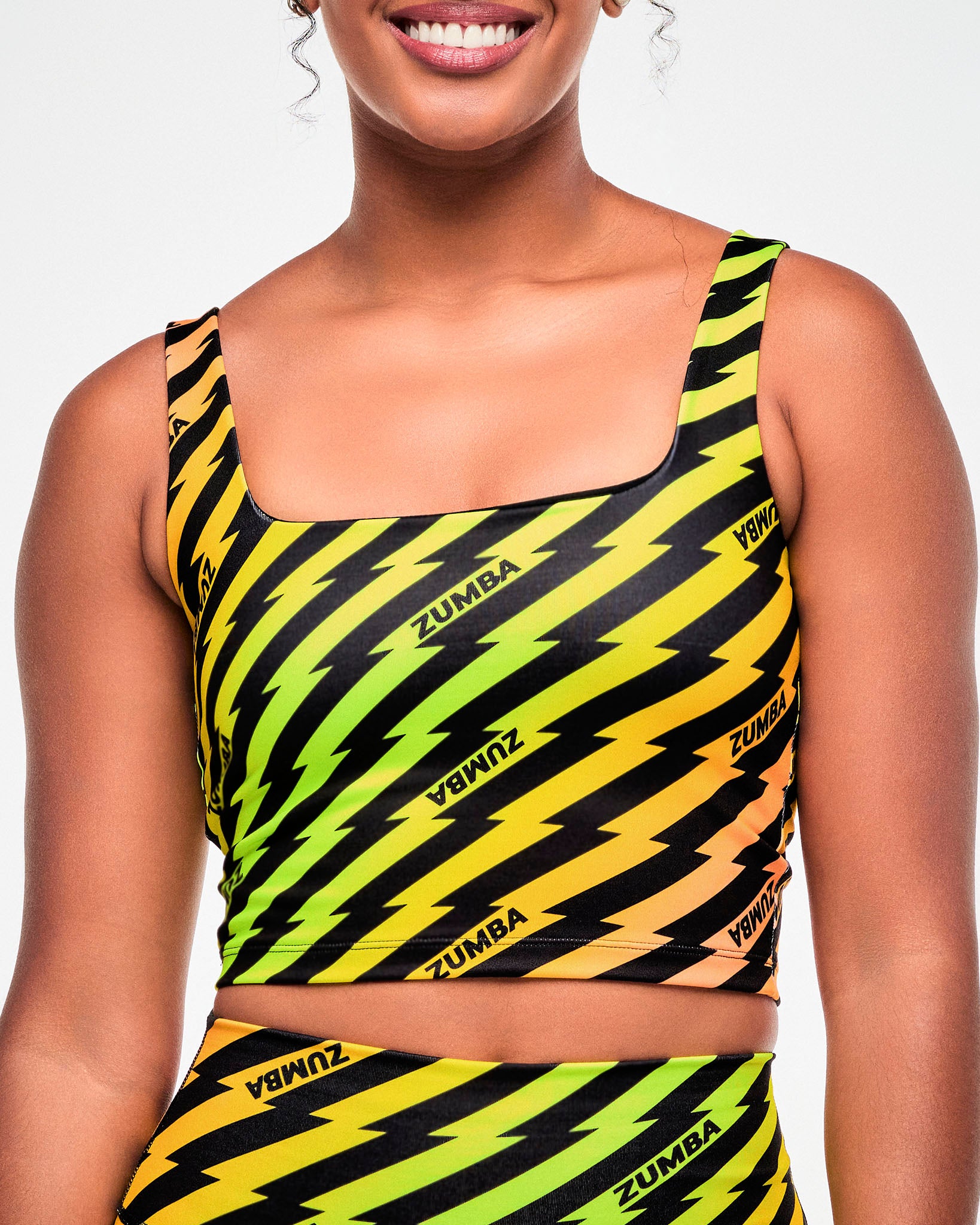 Close-up of a yellow, green, and orange ombre crop tank with shelf bra, with a black lightning bolt zigzag print and repeating small "ZUMBA" text in black and with a square neckline.