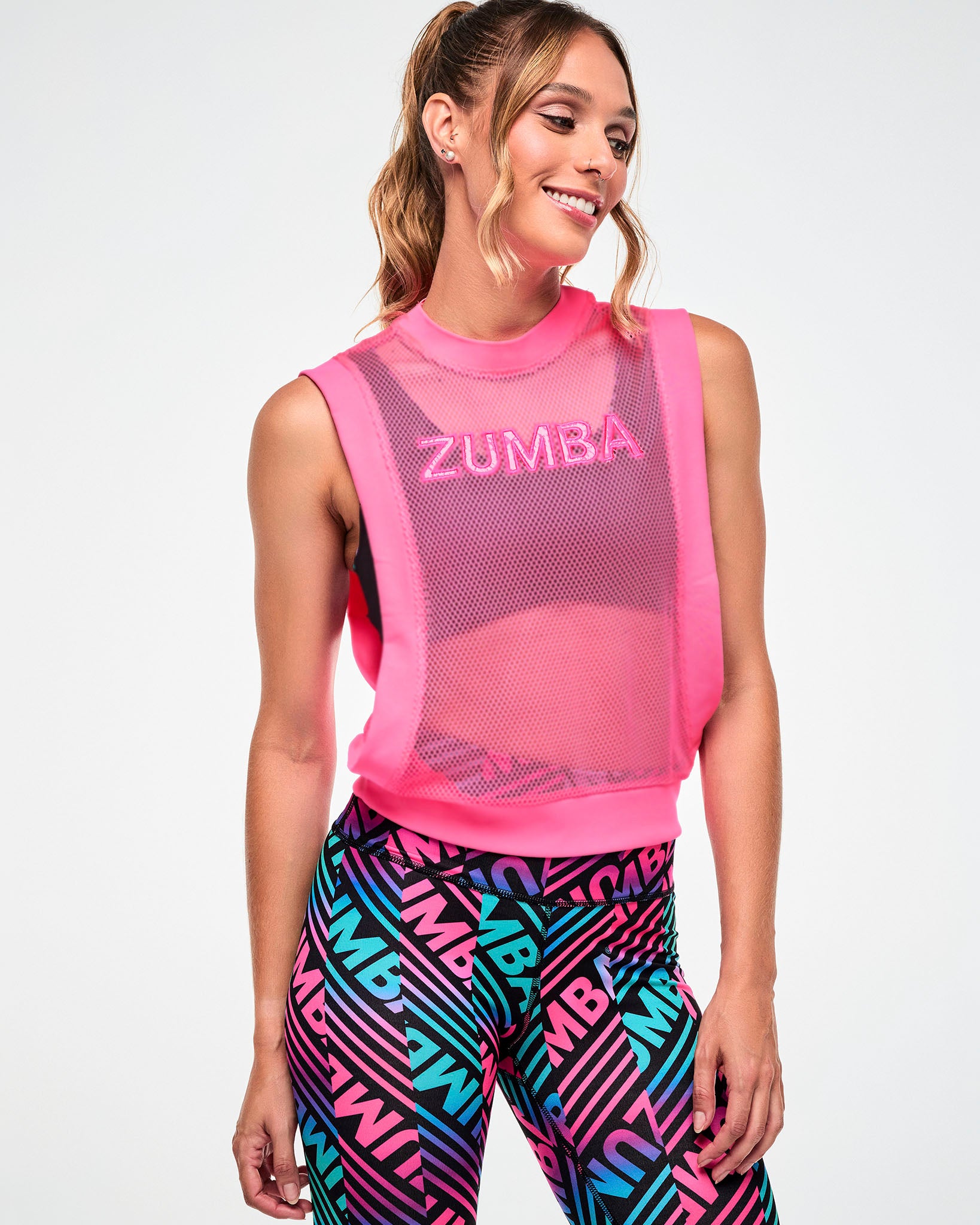 Female model wearing a pink mesh tank with "ZUMBA" text, paired with black, pink, and aqua striped "ZUMBA" print leggings.