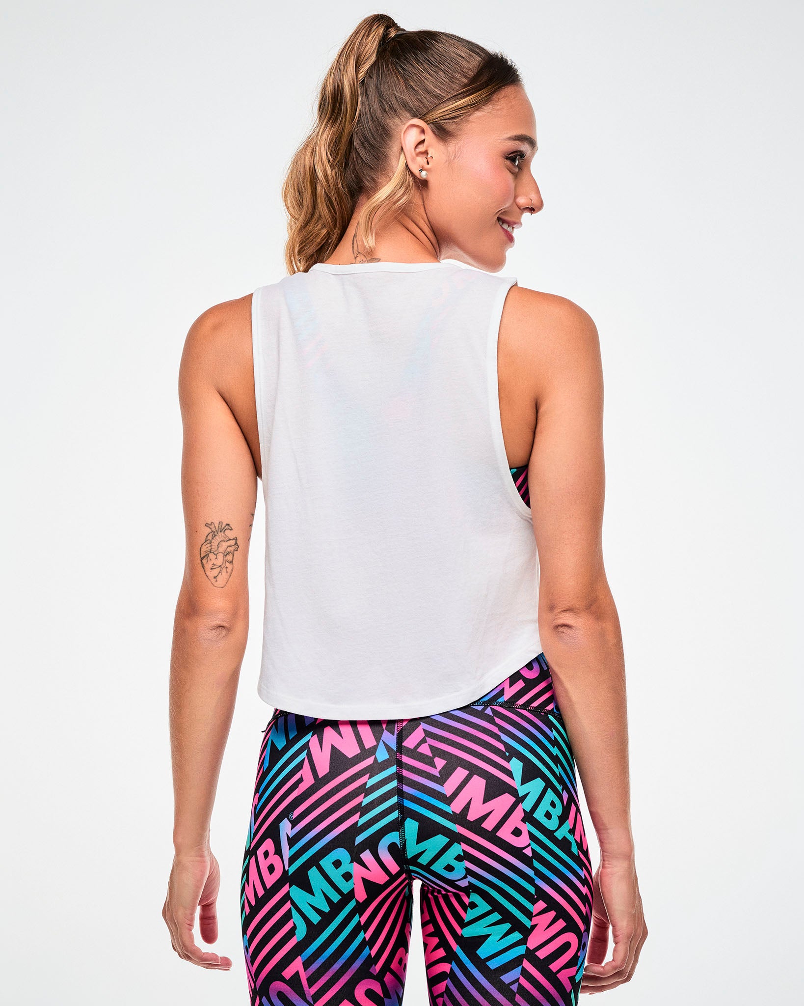 Shown from the back, female model wearing a white cropped tank, paired with blue and pink striped "ZUMBA" print leggings.