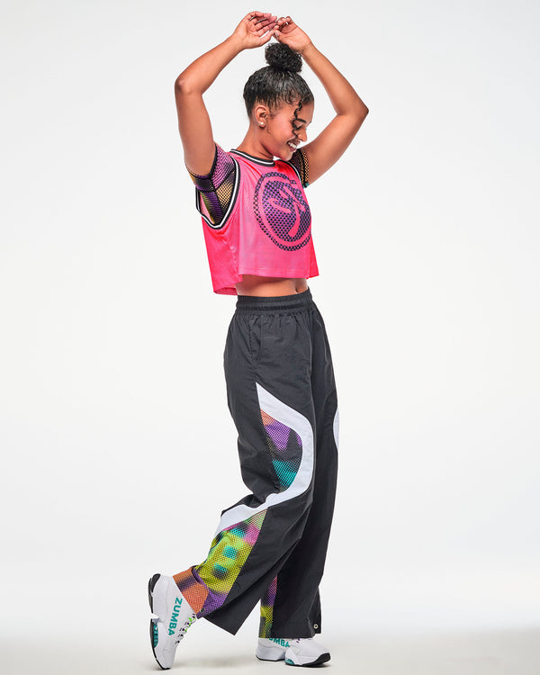 Female model wearing a pink mesh tank, paired with black wide-leg pants with a thick white strip and colorful Zumba print and white shoes.