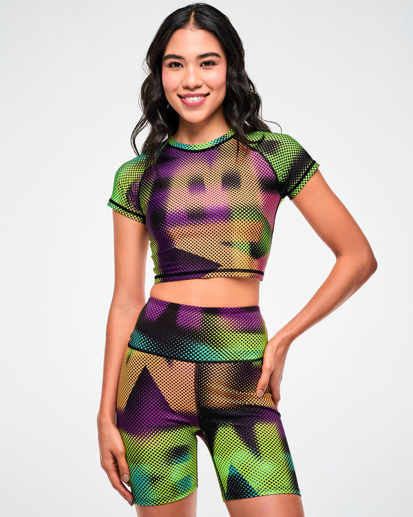 Female model wearing a purple, green, and tan crop top and matching biker shorts in a blurred, oversized 