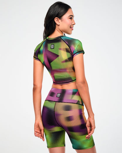 Female model wearing a purple, green, and tan crop top and matching biker shorts in a blurred, oversized "ZUMBA" wordmark print, shown from the back.