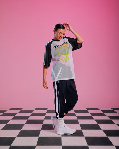 Female model wearing a white mesh raglan tee with black and white striped sleeves, a big yellow and orange Zumbito logo and "ZUMBA" text in black and orange above it, paired with black and white striped joggers with white sneakers.