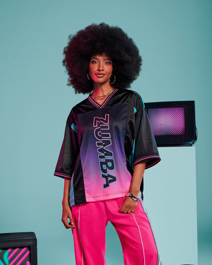 Female model wearing a black jersey tee with an aqua side panel and pink-to-purple gradient "ZUMBA" print, paired with pink track pants, posing against an '80s style blue background with TVs.