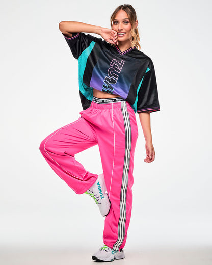 Female model wearing an oversized black jersey tee with an aqua side panel and pink-to-purple gradient "ZUMBA" print, paired with pink track pants with a black and white stripe and white sneakers.