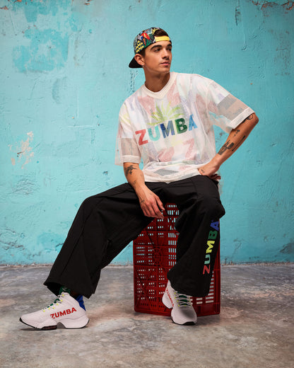 White mesh short-sleeve V-neck top with multicolor “ZUMBA” graphic, modeled by a male wearing black wide-leg pants, seated on a red crate against a turquoise wall with a multicolor backwards cap and white sneakers.