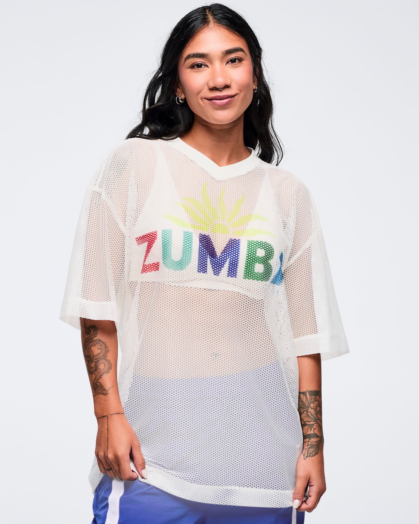 White mesh short-sleeve V-neck top with multicolor “ZUMBA” graphic and loose fit, modeled by a female wearing blue pants on a white studio background.