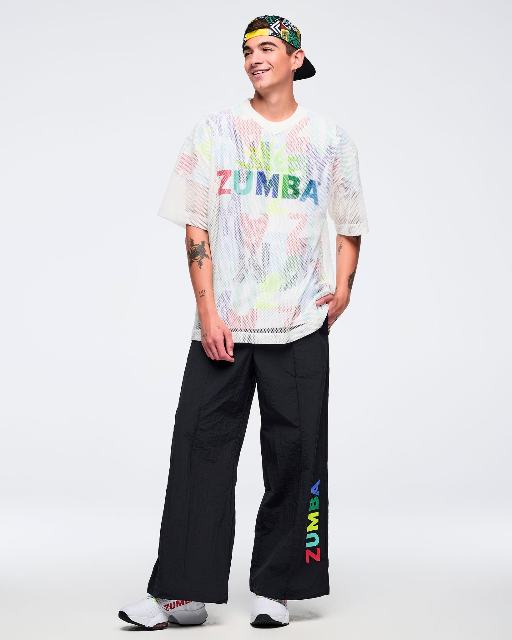 White mesh short-sleeve V-neck top with multicolor “ZUMBA” graphic, modeled by a male wearing black wide-leg pants with a multicolor “ZUMBA” leg print, a multicolor backwards cap, and white sneakers on a white studio background.