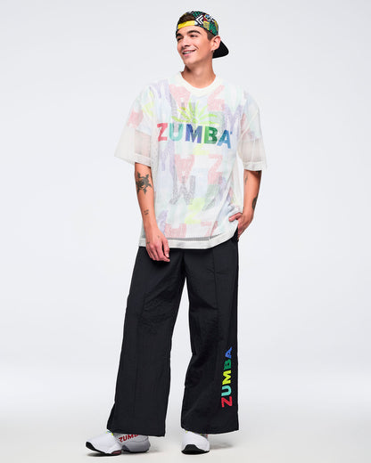 White mesh short-sleeve V-neck top with multicolor “ZUMBA” graphic, modeled by a male wearing black wide-leg pants with a multicolor “ZUMBA” leg print, a multicolor backwards cap, and white sneakers on a white studio background.