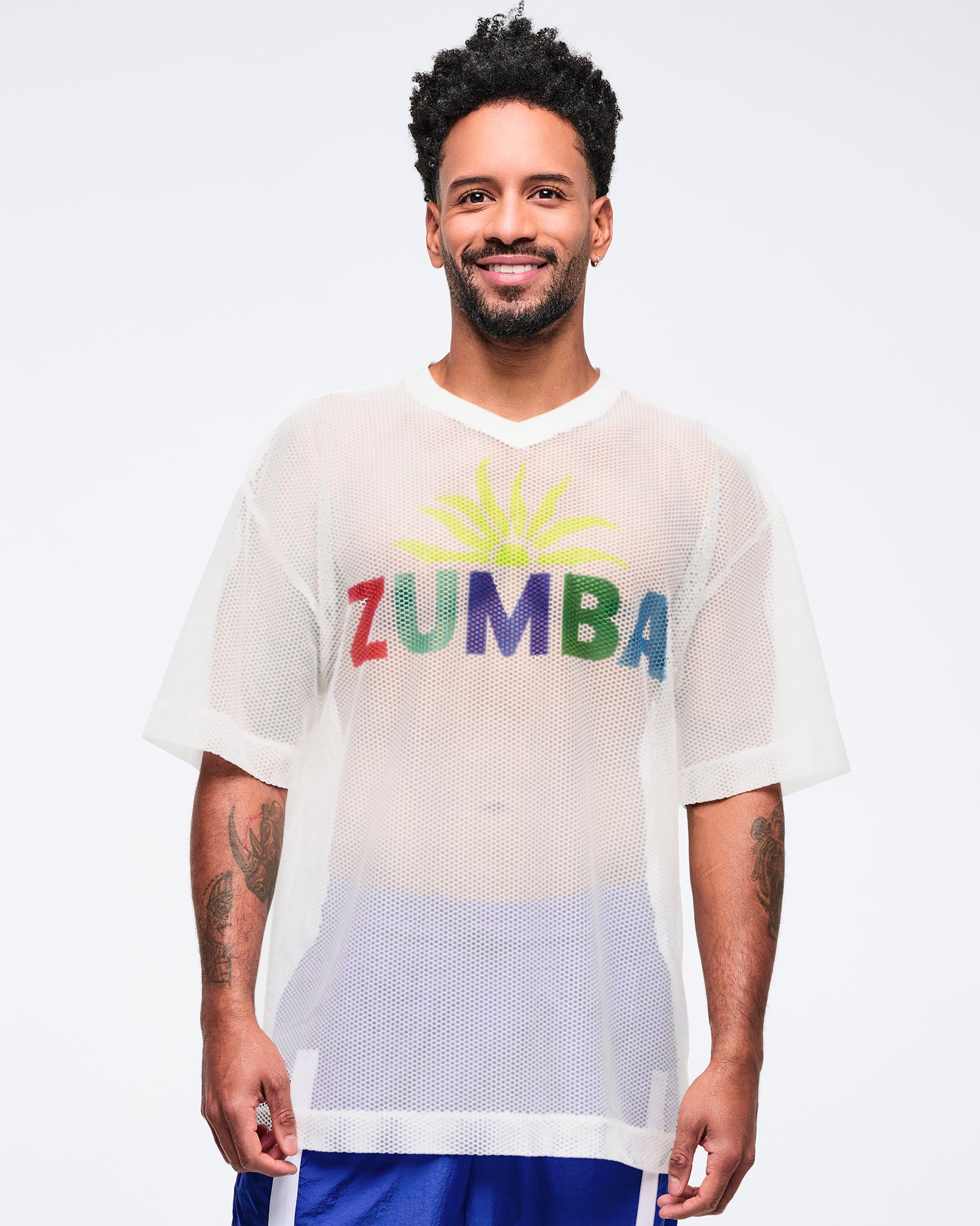 White mesh short-sleeve V-neck top with multicolor “ZUMBA” graphic, modeled by a male wearing blue track pants on a white studio background.