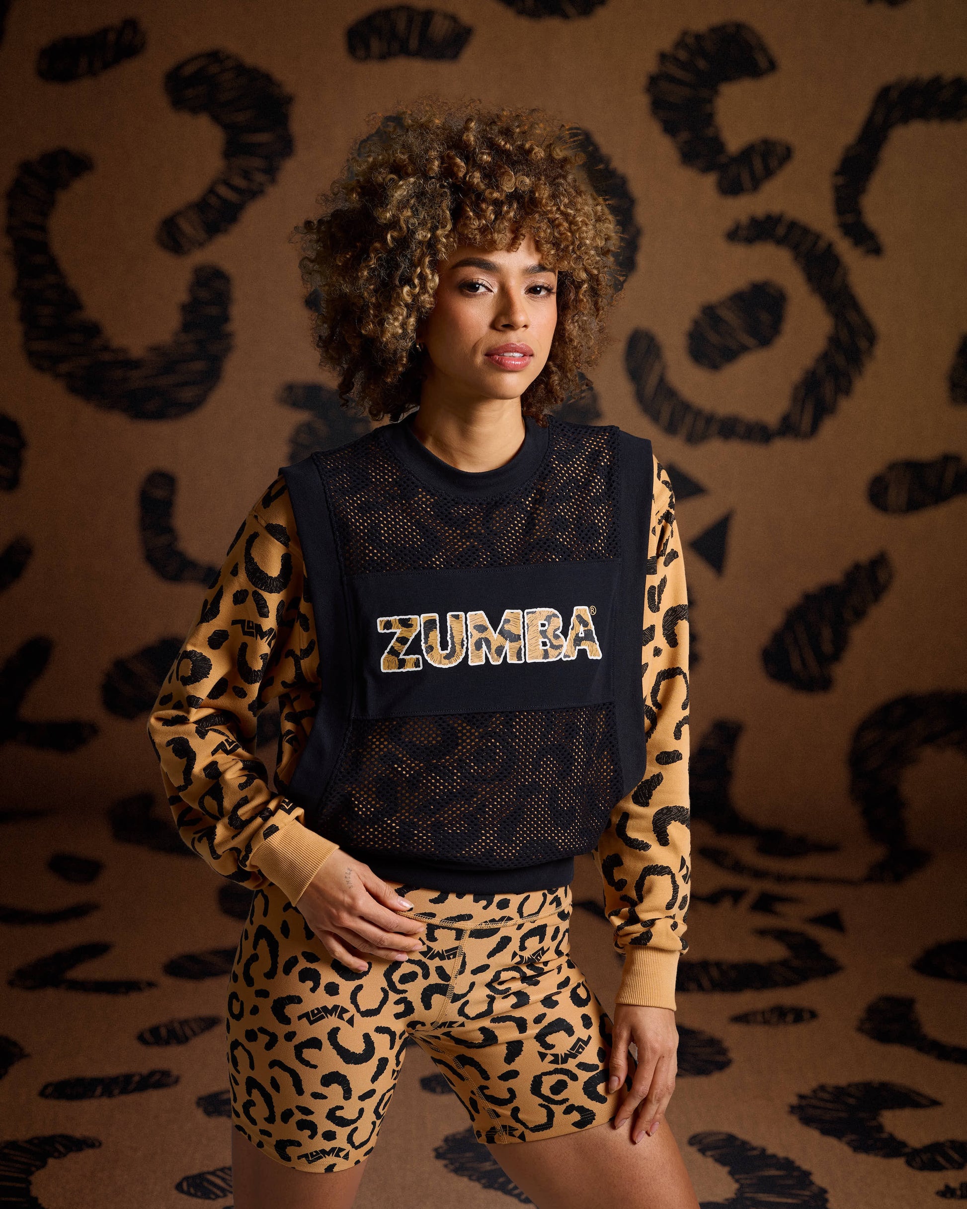 Black mesh Zumba top on female model with a leopard print sweatshirt underneath with biker shorts against a tan leopard backdrop.