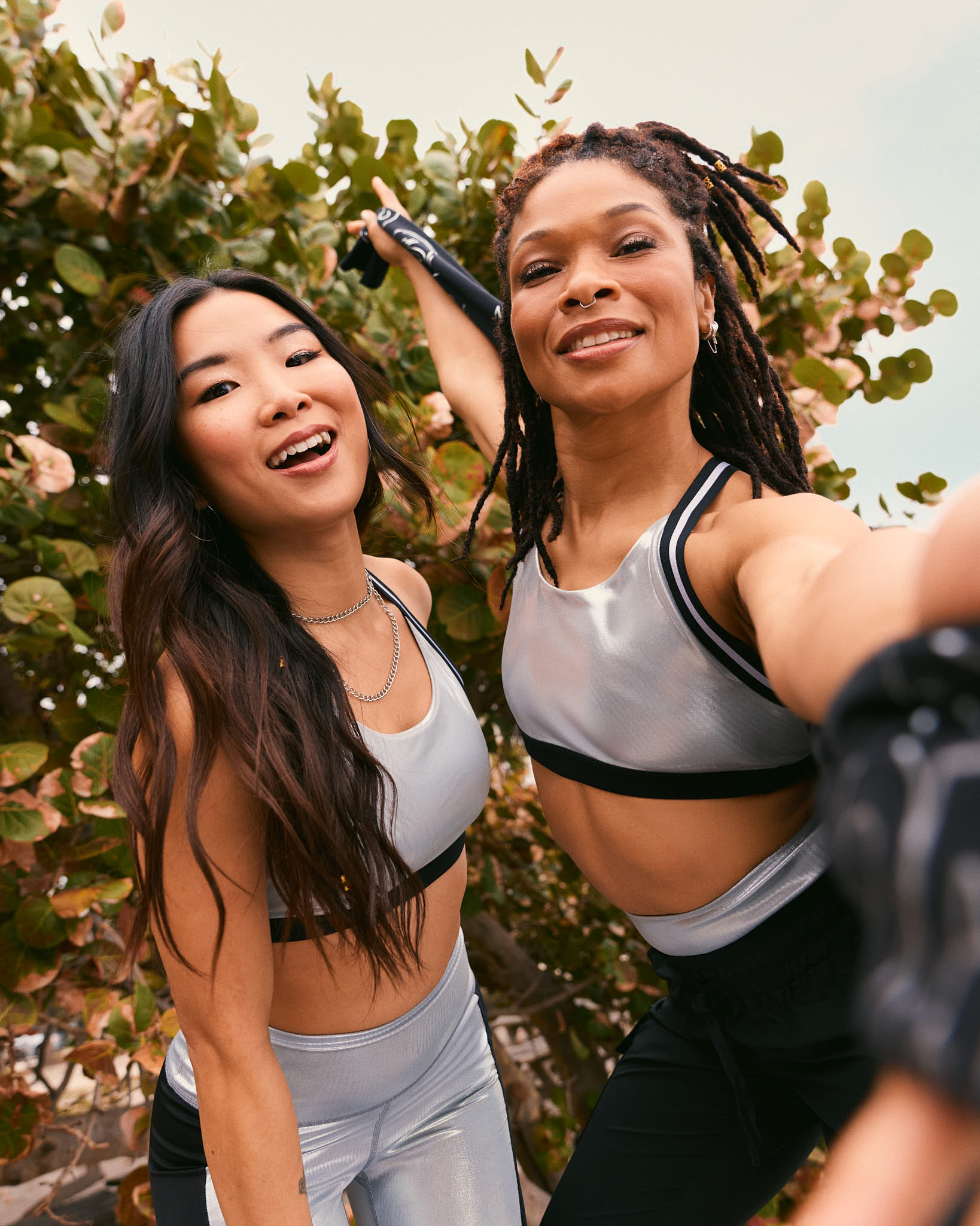 Two female models pose for a selfie outdoors; one wears a silver sports bra with shiny silver leggings, and the other wears a silver sports bra with black jogger pants.