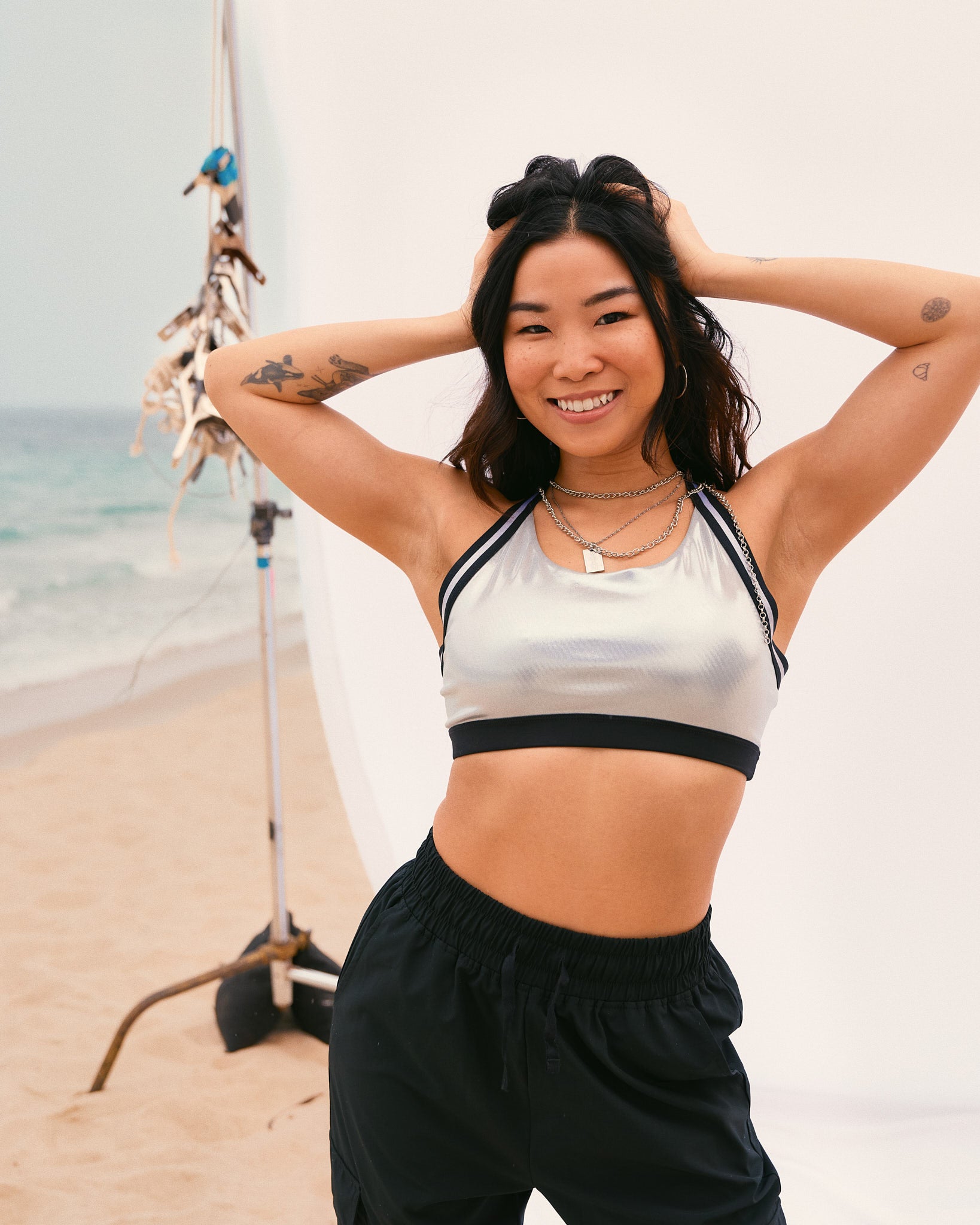 Female model wears the shiny silver cropped sports bra with relaxed black drawstring joggers, smiling with both hands in her hair in front of a white beach backdrop.