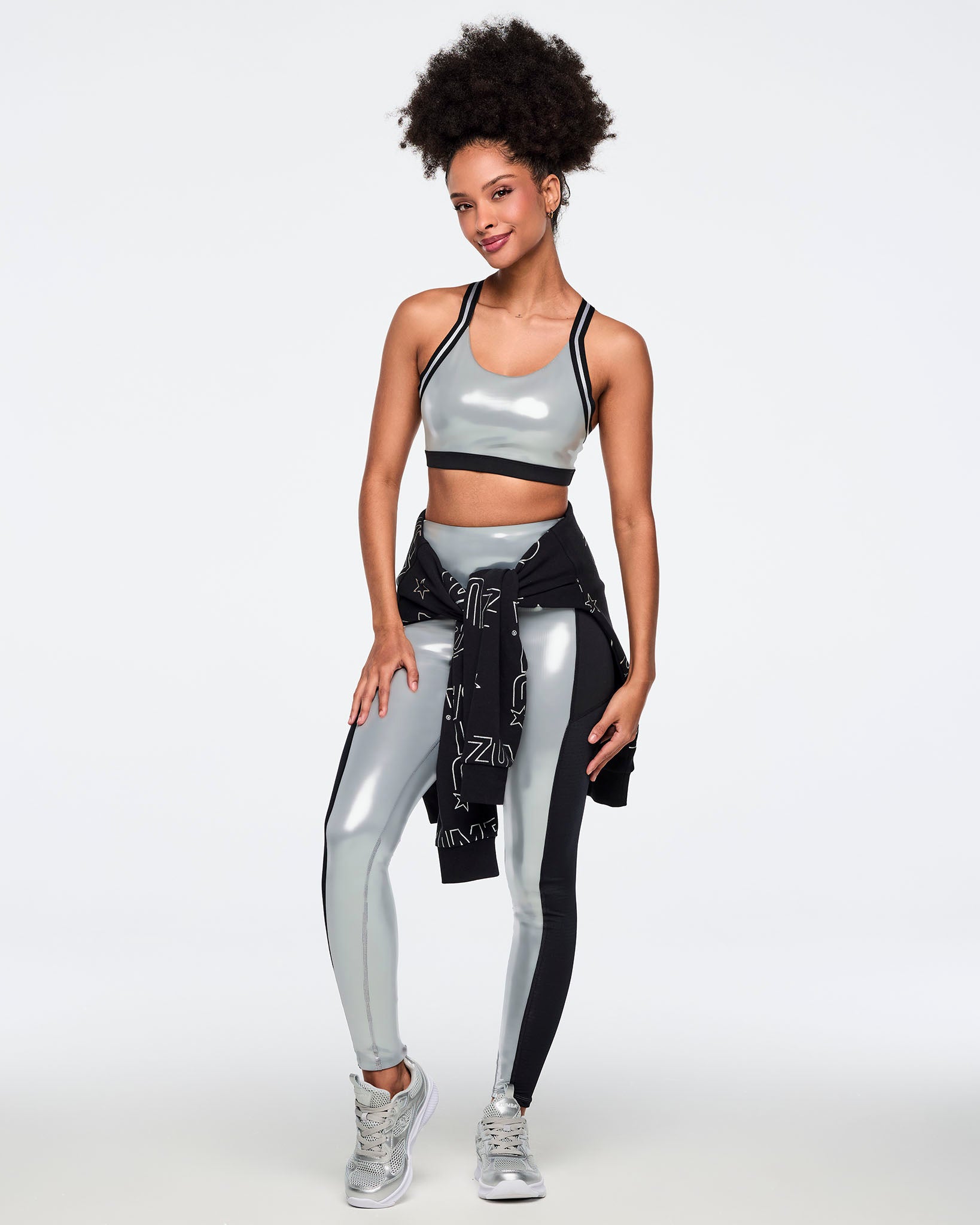 Metallic silver sports bra with black under band and black-and-silver trim straps. Female model wears the glossy bra with matching metallic silver leggings featuring black side panels, a black printed sweatshirt tied at the waist, and silver athletic sneakers, posed in a full-body view against a light gray background.