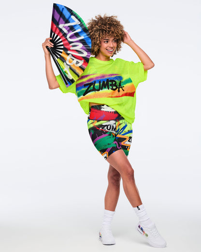 Female model wearing a bright yellow mesh Zumba top with colorful accents, paired with vibrant biker shorts and posing with a rainbow Zumba fan against a white background.
