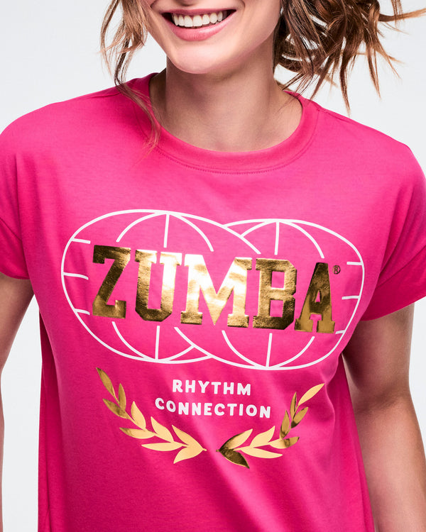 Close-up of a woman in a pink Zumba T-shirt with gold “Zumba” lettering, a gold palm motif, a white globe graphic, and white “Rhythm Connection” text.