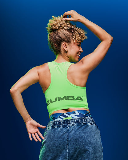 Back view of a female model wearing a neon green crop tank with “Zumba” text along the lower hem, paired with blue denim pants on a dark blue background.