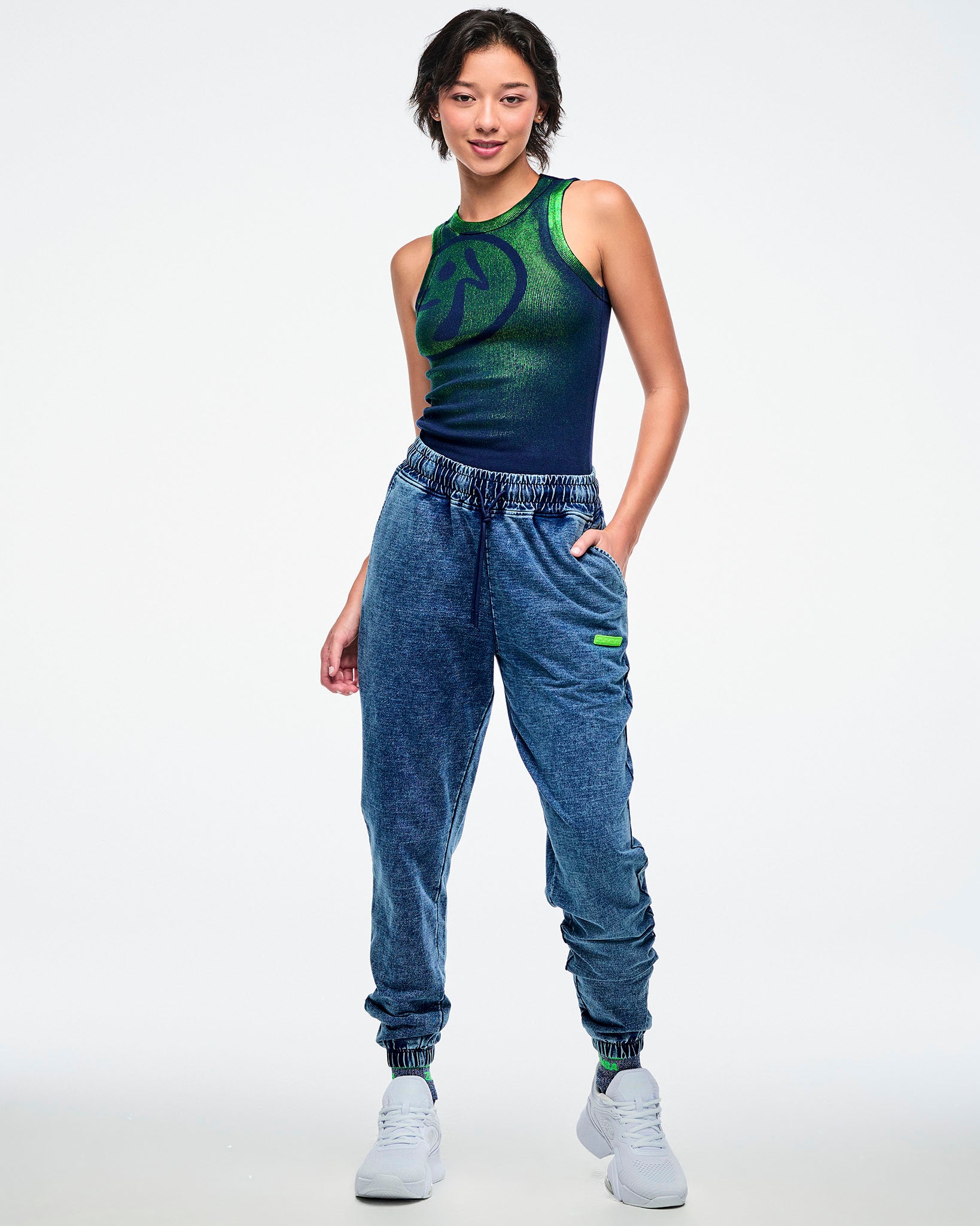 Full view of a female model wearing a green-to-blue gradient tank tucked into blue denim pants and paired with white shoes.