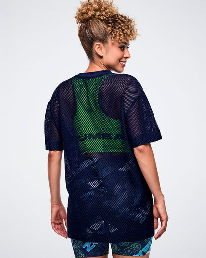 Back view of a female model in an indigo Zumba-branded mesh top layered over a green tank.