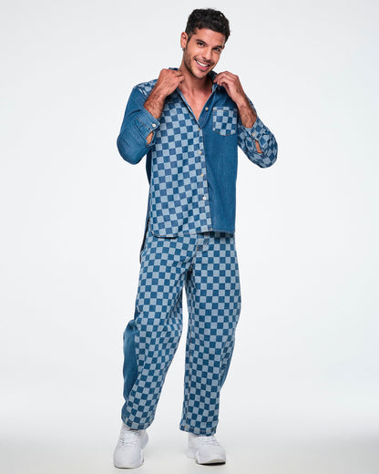 Male model holding his collar in a long-sleeve denim button-up split between a light and medium blue checkerboard right front and a solid medium-wash left front, with a checkerboard chest pocket and one checkerboard sleeve. Paired with checkerboard denim barrel ankle pants.