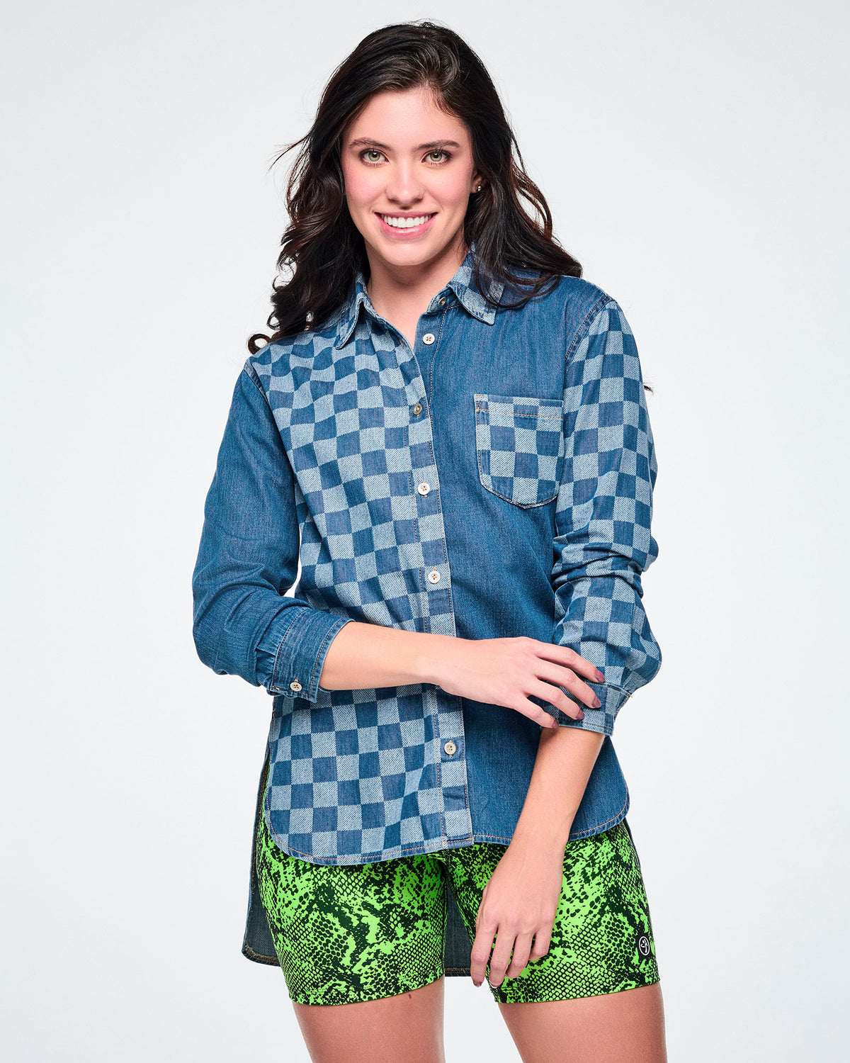 Front view of a female model wearing a denim button-down with a checkerboard front panel, left sleeve, and chest pocket, paired with neon green snake-print biker shorts.