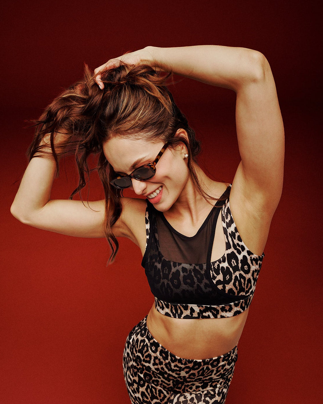 A female model posing in a matching leopard-print activewear set consisting of a sports bra and leggings, paired with white shoes against a solid dark red studio background.