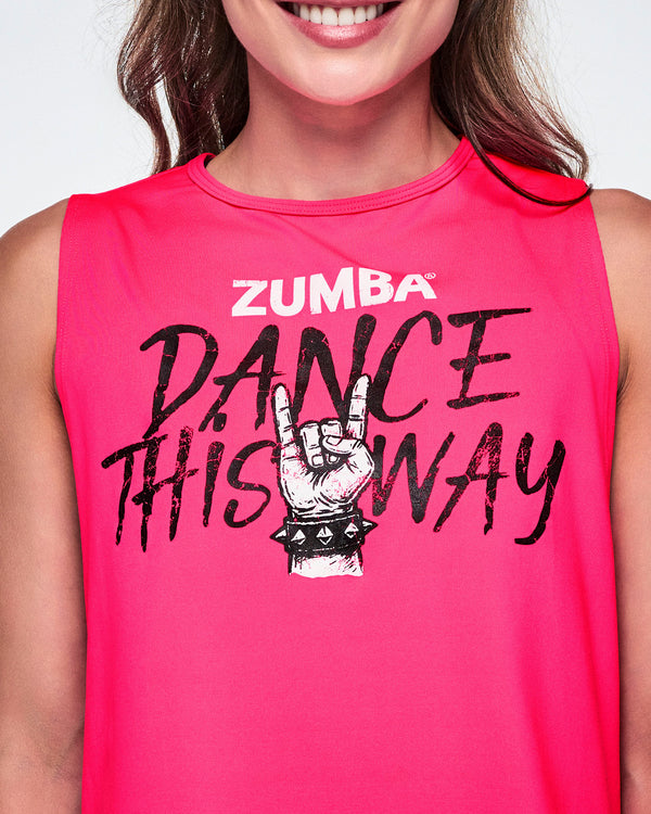 Close-up of a female model wearing a bright pink sleeveless tank top with a Zumba graphic that reads 'Dance This Way' in black lettering, with a hand making a rock gesture and Zumba at the top in white.