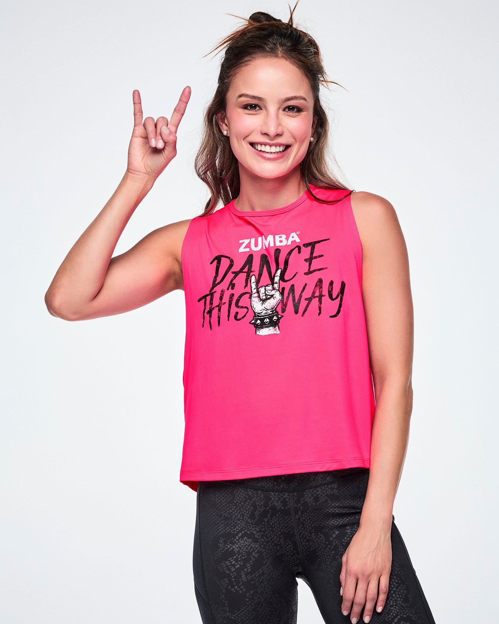 A female model wearing a bright pink sleeveless Zumba tank top that reads 'Dance This Way' in black lettering, with a hand making the same rock gesture as the model, and Zumba at the top in white and paired with black leggings.