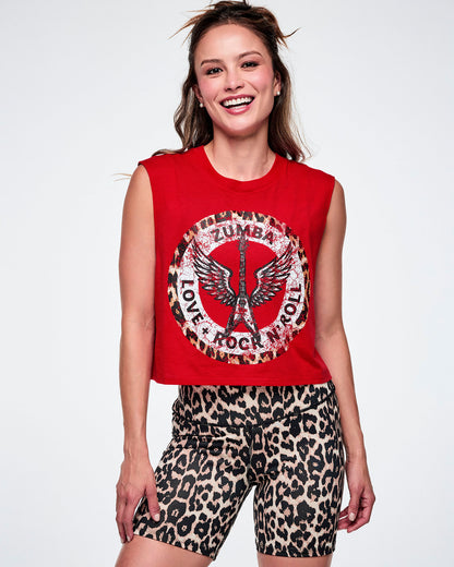 A female model wearing a red sleeveless crop tank with a large white, distressed, circular Zumba rock graphic on the front with a leopard print border. In the circle it says ZUMBA at the top and LOVE + ROCK N' ROLL at the bottom, with a white guitar and wings outlined in the middle. This is paired with tan leopard print biker shorts.
