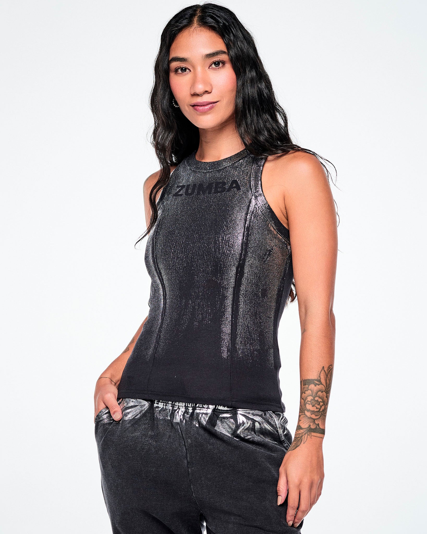 A female model wearing a sleeveless black Zumba ribbed tank top with a subtle metallic sheen and 'ZUMBA' printed across the chest, in smaller black text, paired with dark pants, against a plain white studio background.