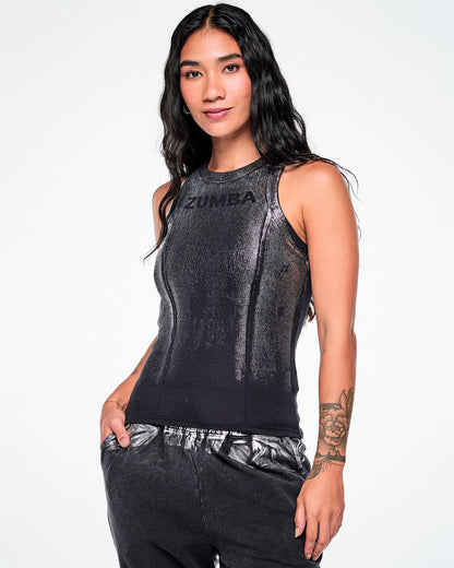 A female model wearing a sleeveless black Zumba ribbed tank top with a subtle metallic sheen and 'ZUMBA' printed across the chest, in smaller black text, paired with dark pants, against a plain white studio background.