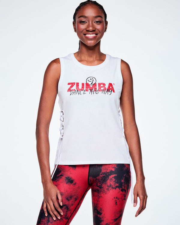 Front view of a female model wearing a white sleeveless tank top with a red ZUMBA text and overlayed with a small black Zumbito on top and on the bottom, black cursive text saying 