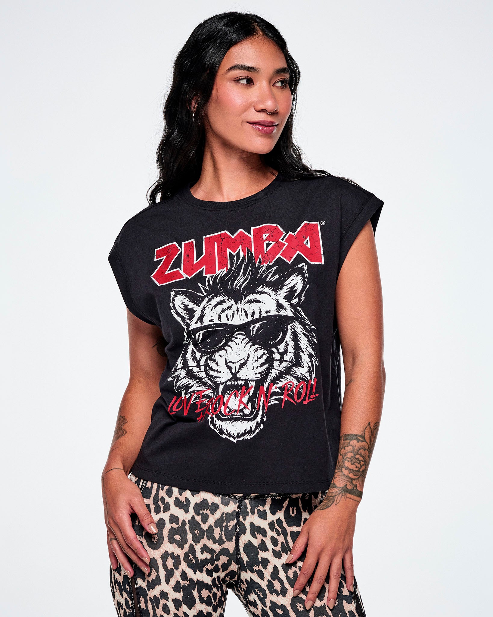 A female model wearing a black tank with a large graphic print of a black and white tiger wearing sunglasses and the word 'ZUMBA' in red rock-like text on the top and "LOVE + ROCK N' ROLL", over the tigers mouth, all paired with leopard print leggings.
