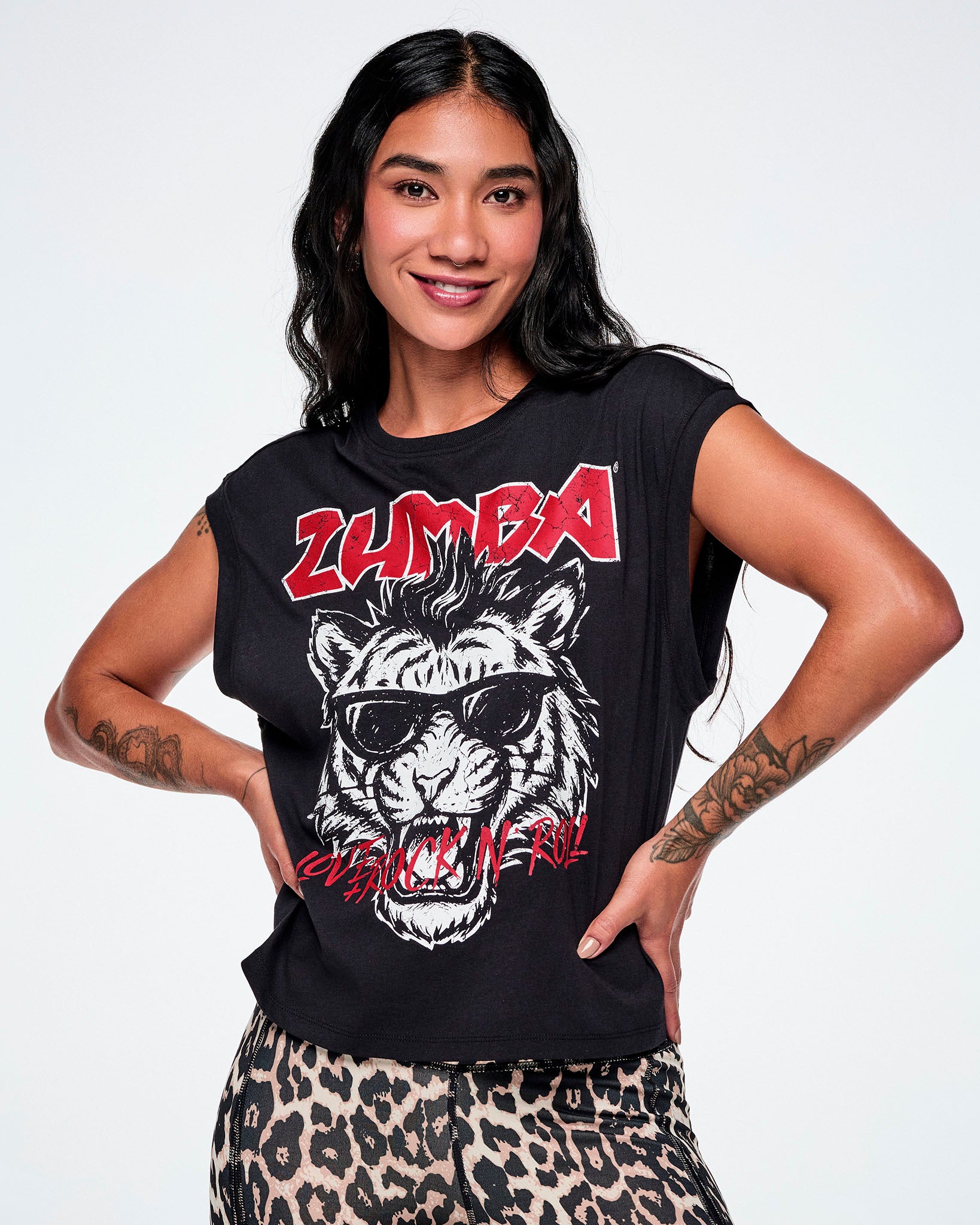 A female model with hands on her hips, wearing a black tank with a large graphic print of a black and white tiger wearing sunglasses and the word 'ZUMBA' in red rock-like text on the top and "LOVE + ROCK N' ROLL", over the tigers mouth, all paired with leopard print leggings.