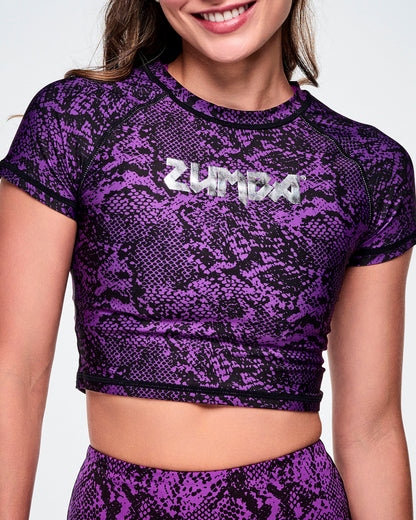 A close-up of a female model wearing a purple and black snakeskin-patterned short-sleeve crop top with the ZUMBA rock inspired text, printed in silver across the chest.