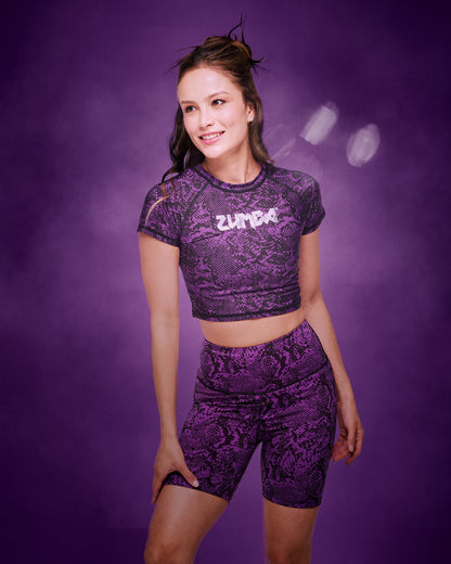 A female model wearing a matching purple and black snakeskin-patterned activewear set consisting of a short-sleeve crop top with 'ZUMBA' printed on the chest, in silver and matching high-waisted bike shorts, set against a smoky purple studio background.