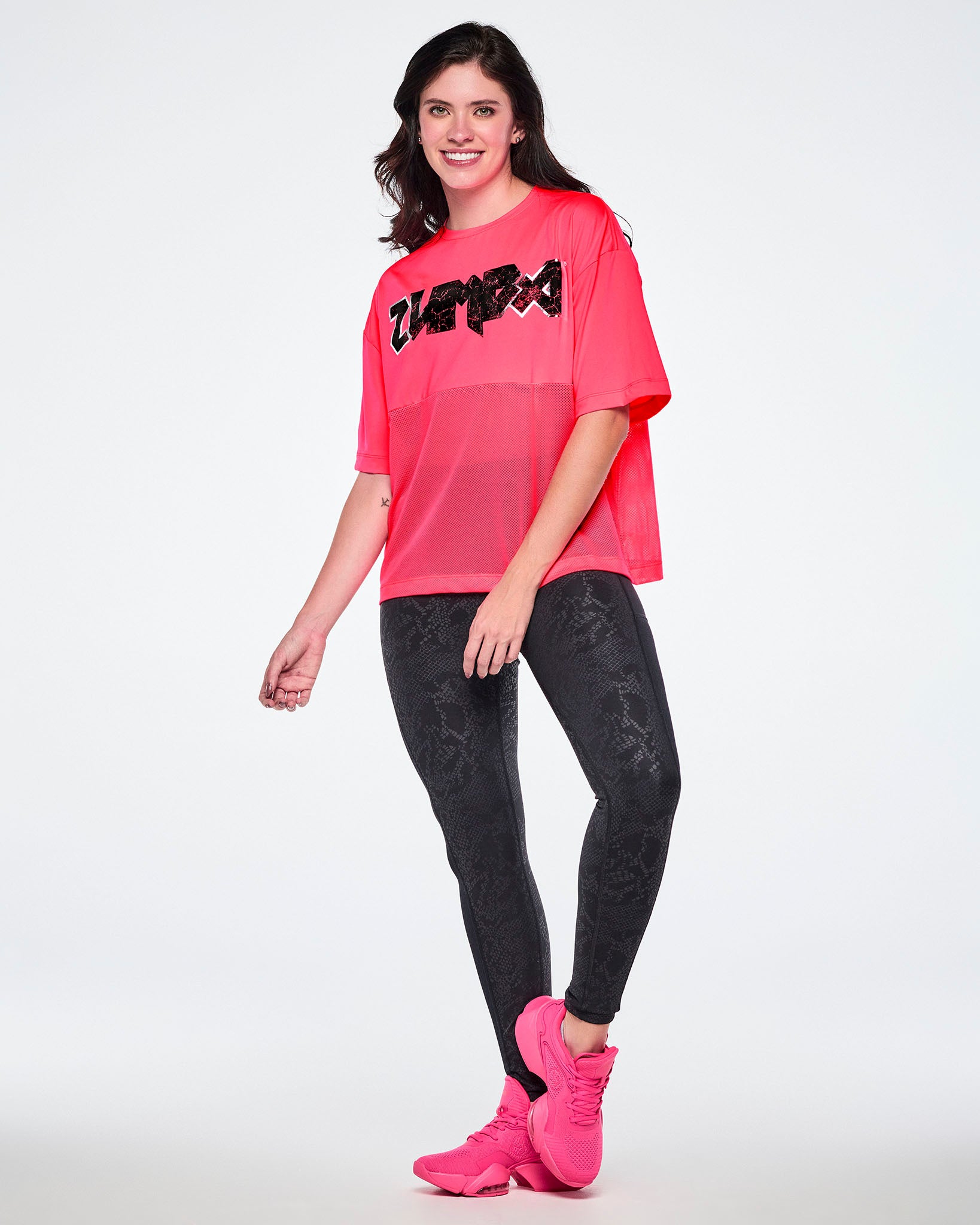 Full view of a female model wearing a bright pink short-sleeve top with mesh bottom panels and black and silver distressed 'ZUMBA' rock-like text across the chest, paired with black leggings and pink shoes.