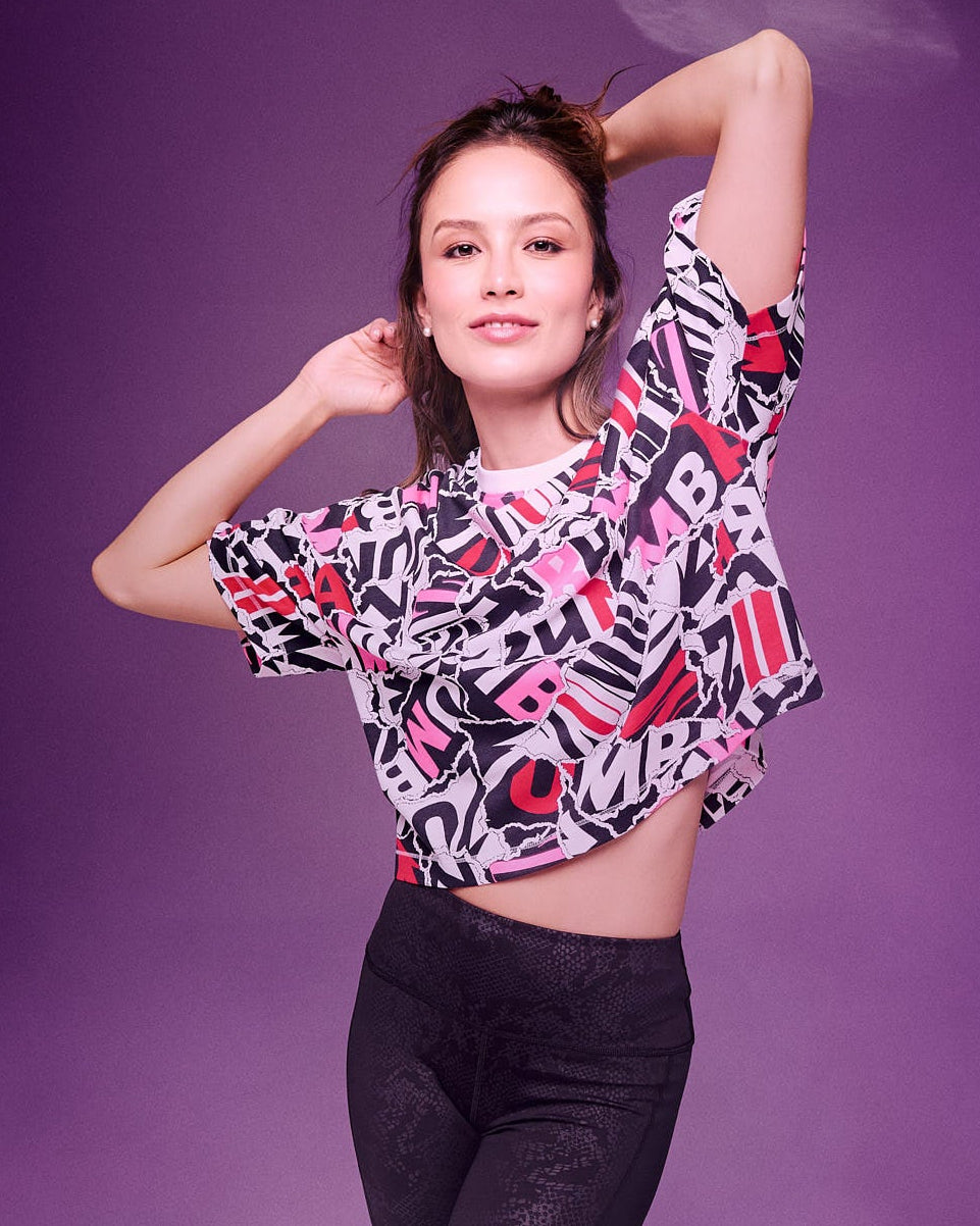A female model posing in a vibrant studio setting against a purple background, wearing a short-sleeve crop top with a colorful abstract ZUMBA text pattern in pink, red, black, and white, paired with black leggings.