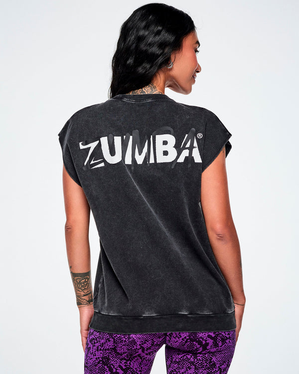 A female model shown from the back wearing a faded black Zumba top with large white ZUMBA text, with a light grey ZUMBA overtop this, all across the upper back and paired with purple snake-skin patterned biker shorts.