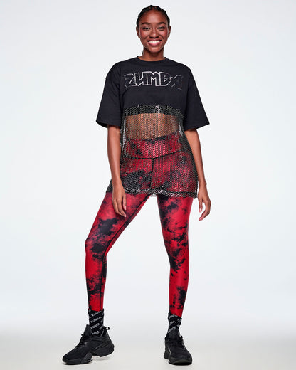 Full view of a female model wearing a black Zumba cropped top with a sheer mesh panel with sequins and a large silver outlined ZUMBA logo across the chest, paired with red and black leggings, black socks and shoes.