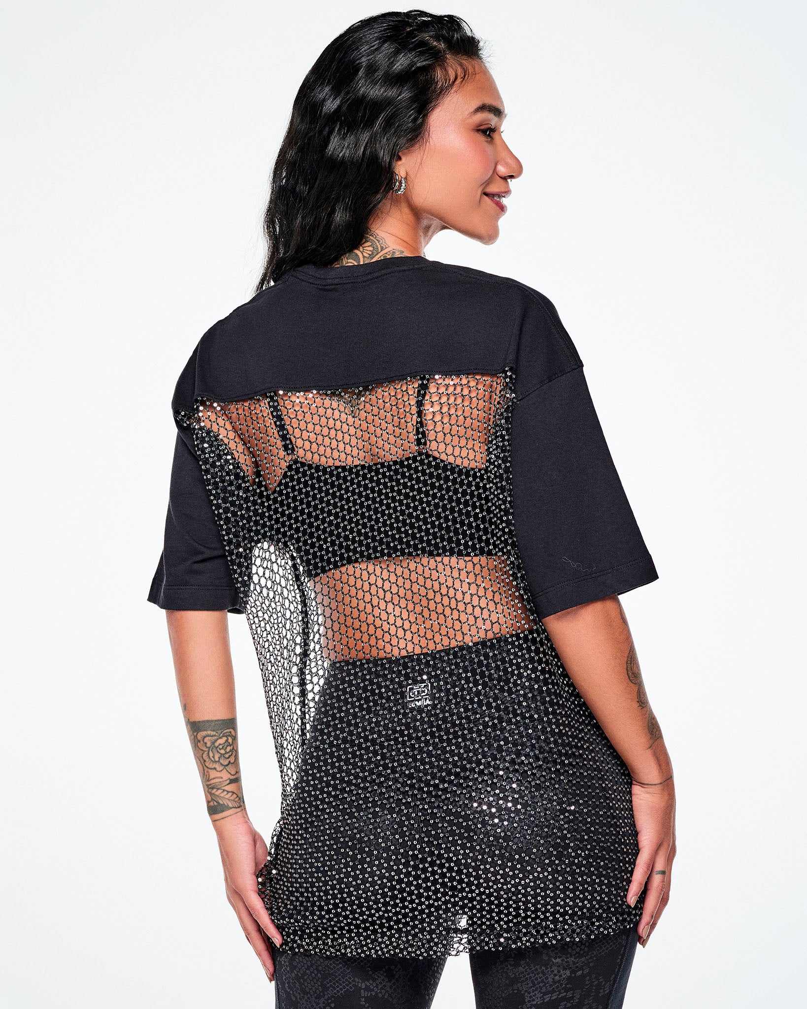 A female model shown from the back wearing a black tee, with a larger mesh and chain-link sheer mesh panel with sequins covering most of her back, paired with black leggings.